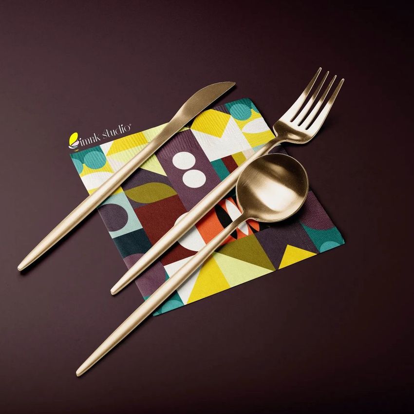 Paper napkin mockup with a colorful geometric pattern, placed on a dark surface with gold cutlery arranged on top.