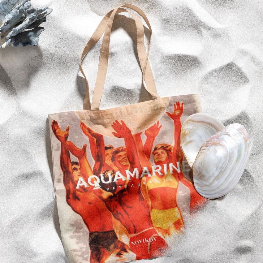 Tote bag mockup on light sand with large seashells, featuring a vintage-style print of people with raised arms and bold text overlay.