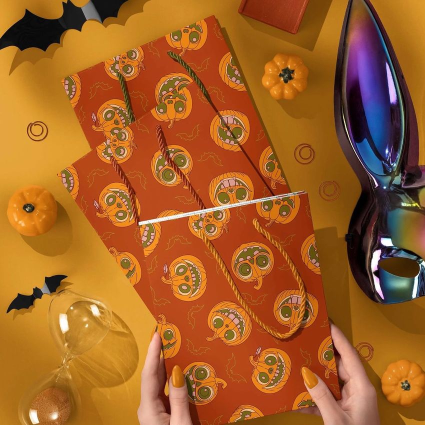 Halloween paper gift bag mockup with orange pumpkin faces pattern, held by hands on a yellow background with festive props.
