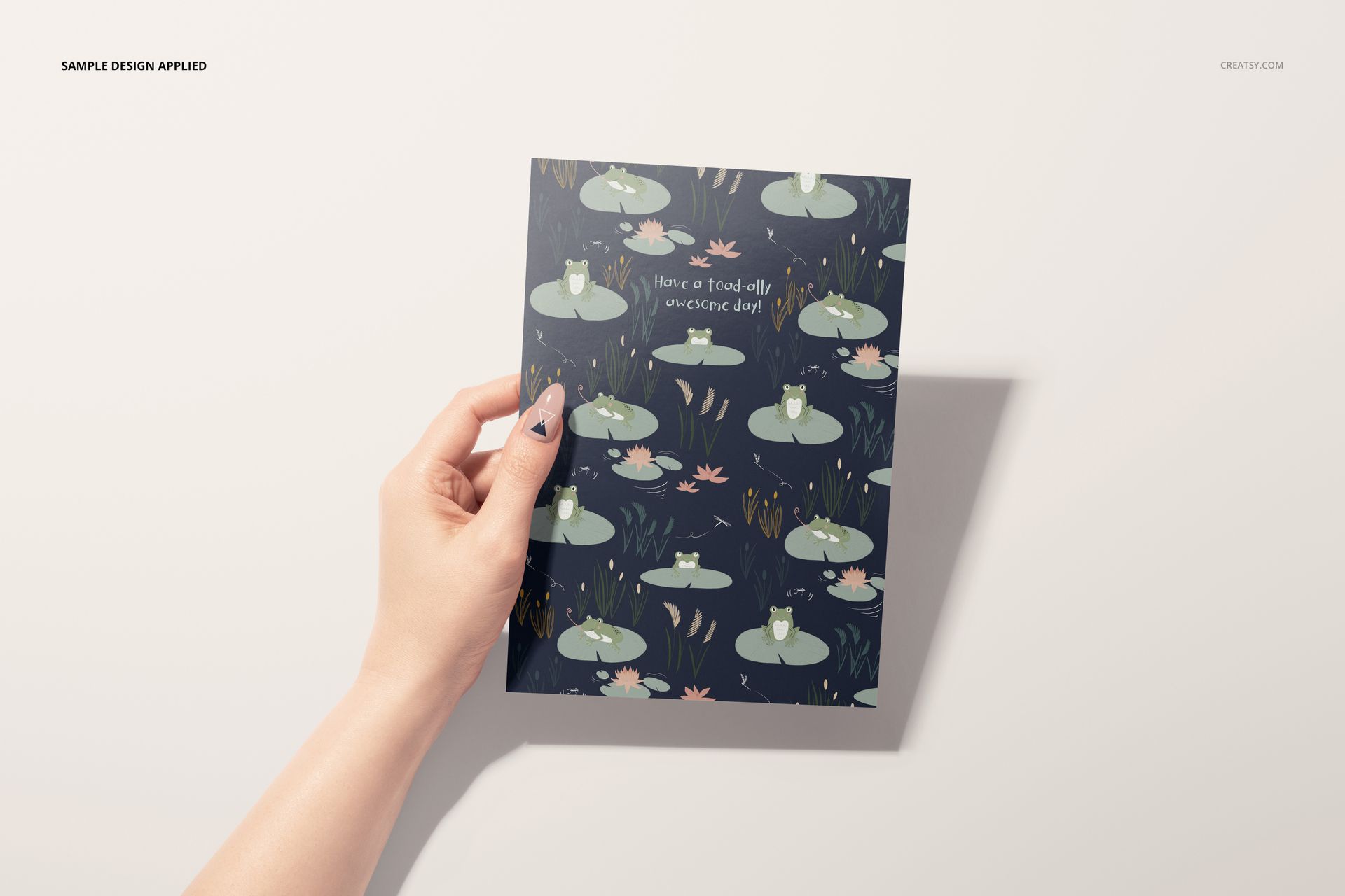 Close-up of a female hand displaying a greeting card featuring whimsical frogs and lily pads, stationery mockup scene.