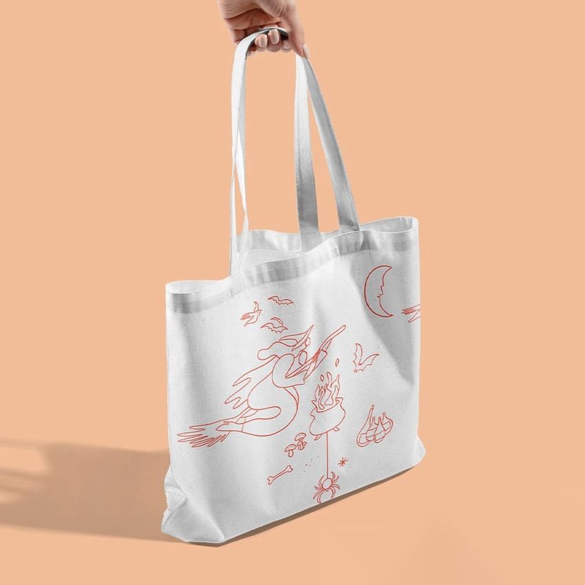 White tote bag mockup with orange line art illustrations of a witch, moon, and Halloween-themed icons, held by a hand on a peach background.