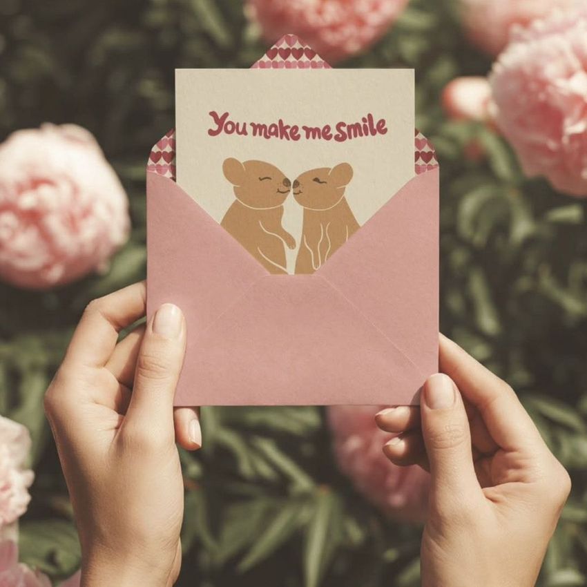 A greeting card mockup with two cartoon animals and text inside a pink envelope, held by hands in front of pink peony flowers.