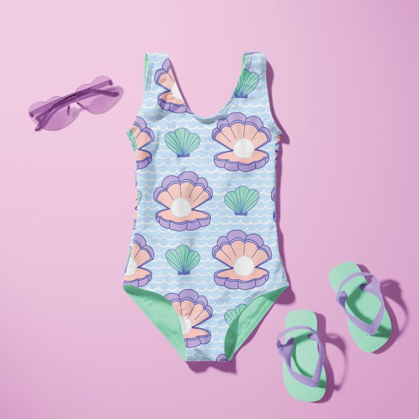 Baby swimsuit mockup featuring a shell pattern in pastel colors, displayed flat on a pink background with sunglasses and sandals.