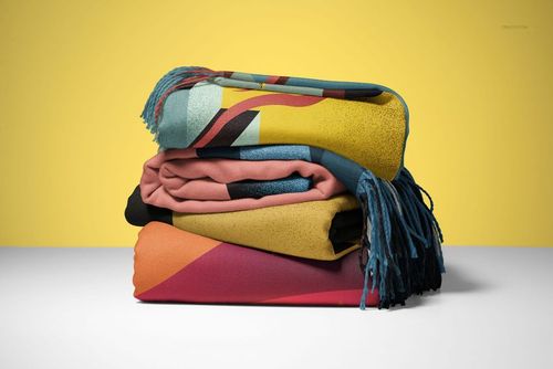 PSD mockup of folded tassel fringe blankets with colorful geometric prints, displayed on a white tabletop with a yellow wall.