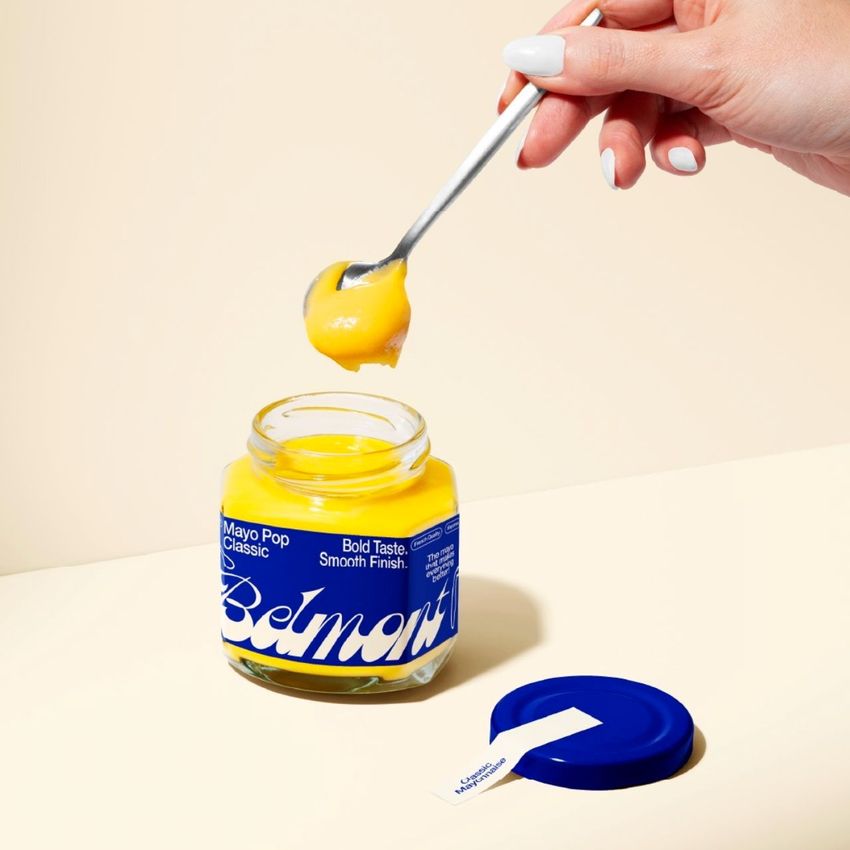 Glass jar mockup with bright yellow mango jam and a bold blue label, shown with a spoon and detached blue lid on a beige background.