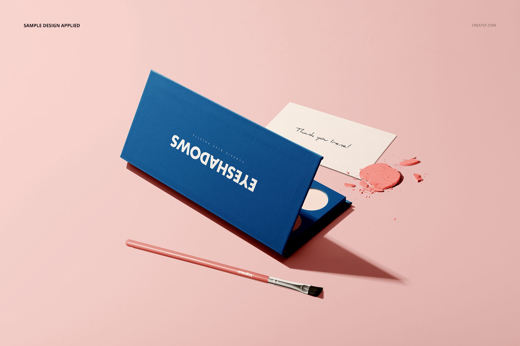 Blue eyeshadow palette mockup shown on a light pink background with a makeup brush and scattered powder detail.