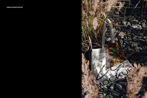 Tote bag mockup resting on dark rocks among tall dry grass, featuring white handles and a customizable design area.