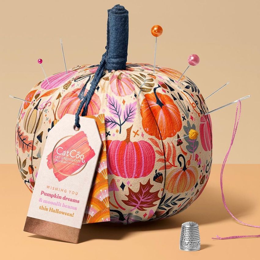 Fabric pumpkin mockup decorated with colorful autumn patterns, used as a pincushion with pins and a tag attached, set on a beige surface.