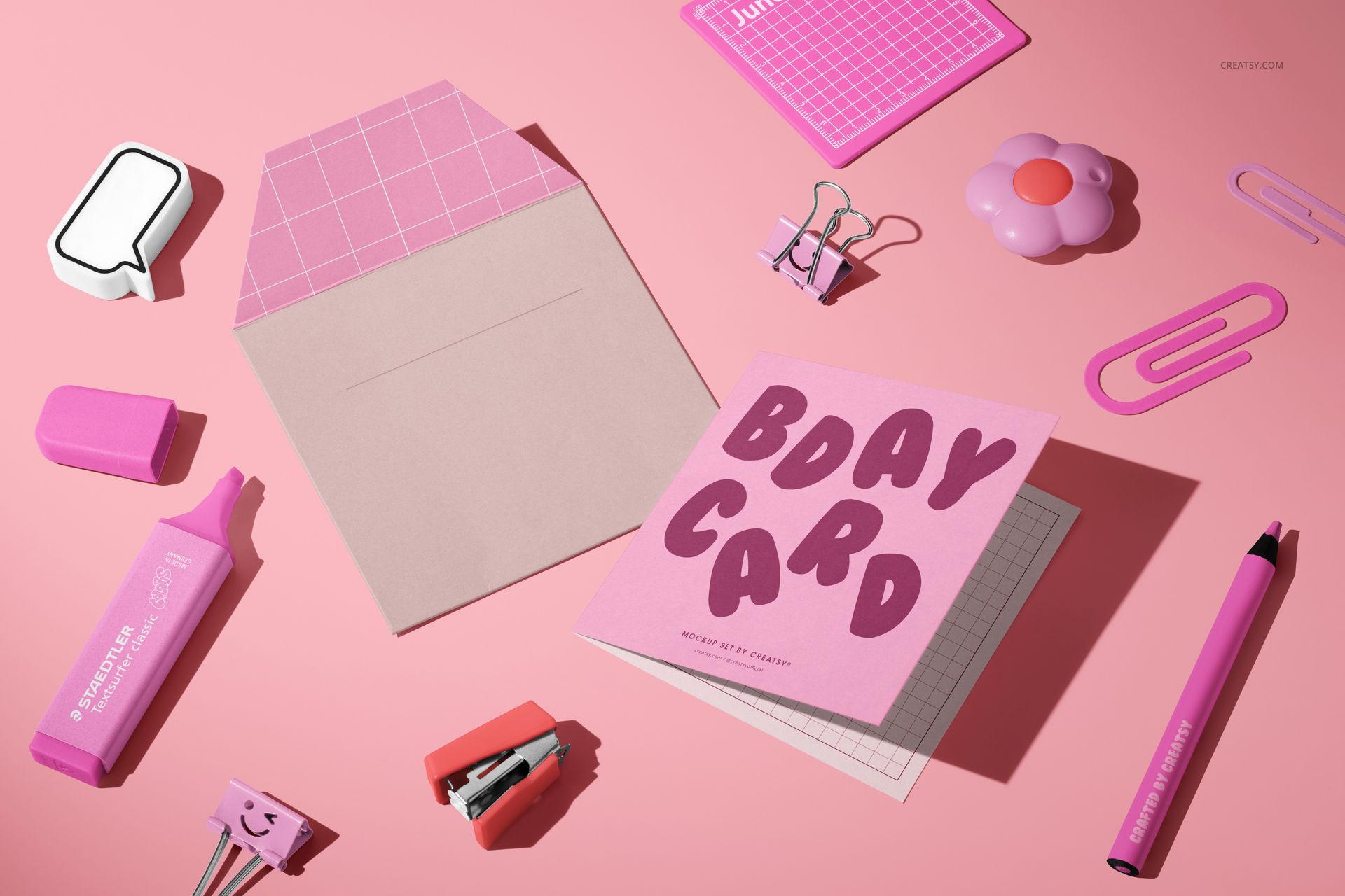 PSD mockup of a square greeting card and envelope on a pink background with various office supplies nearby.