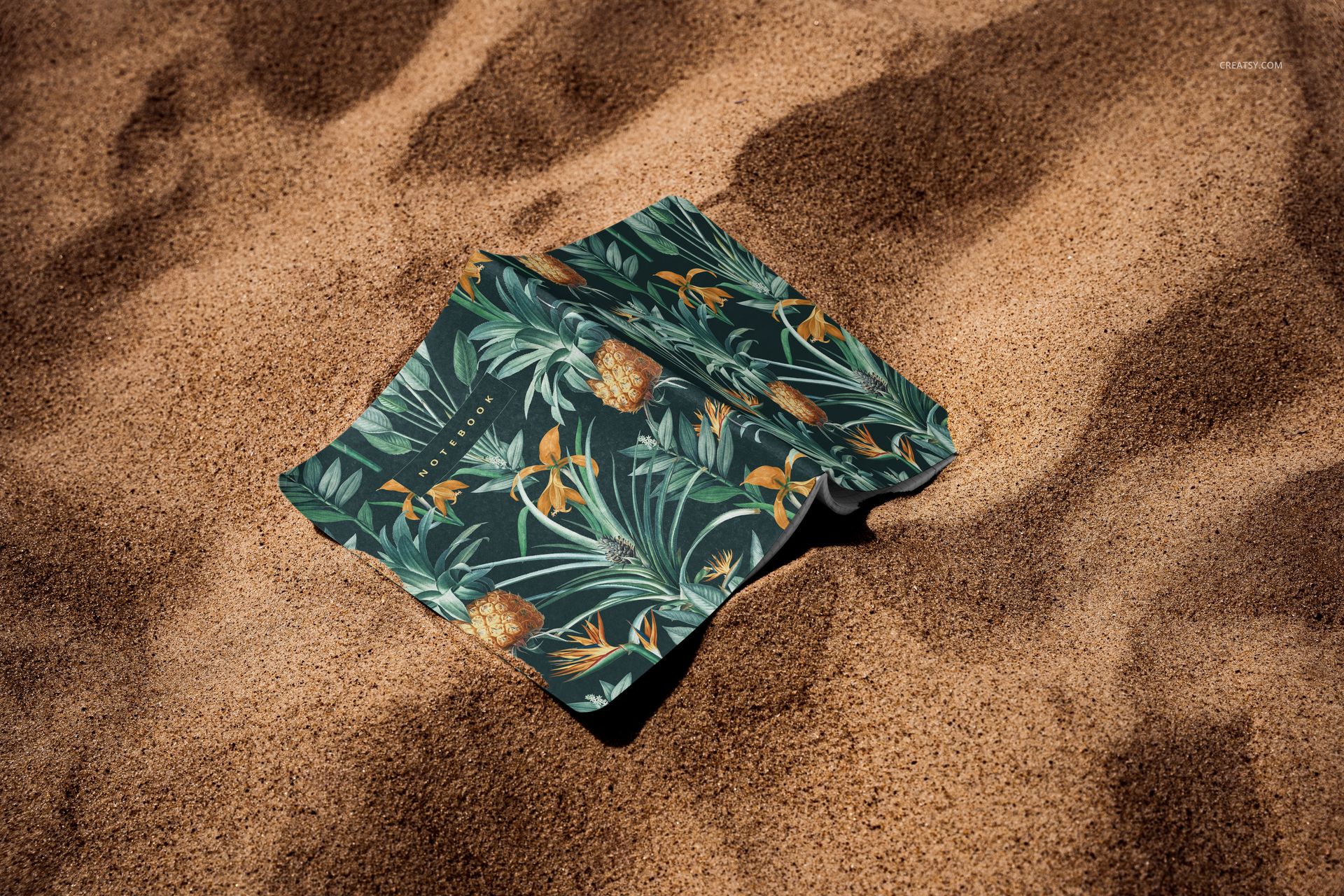 PSD mockup of a stationery notebook with botanical cover resting on beach sand, overhead view.