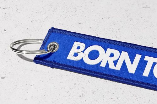 Blue rectangular keychain mockup featuring white lettering and a silver ring, displayed on a concrete-like background.