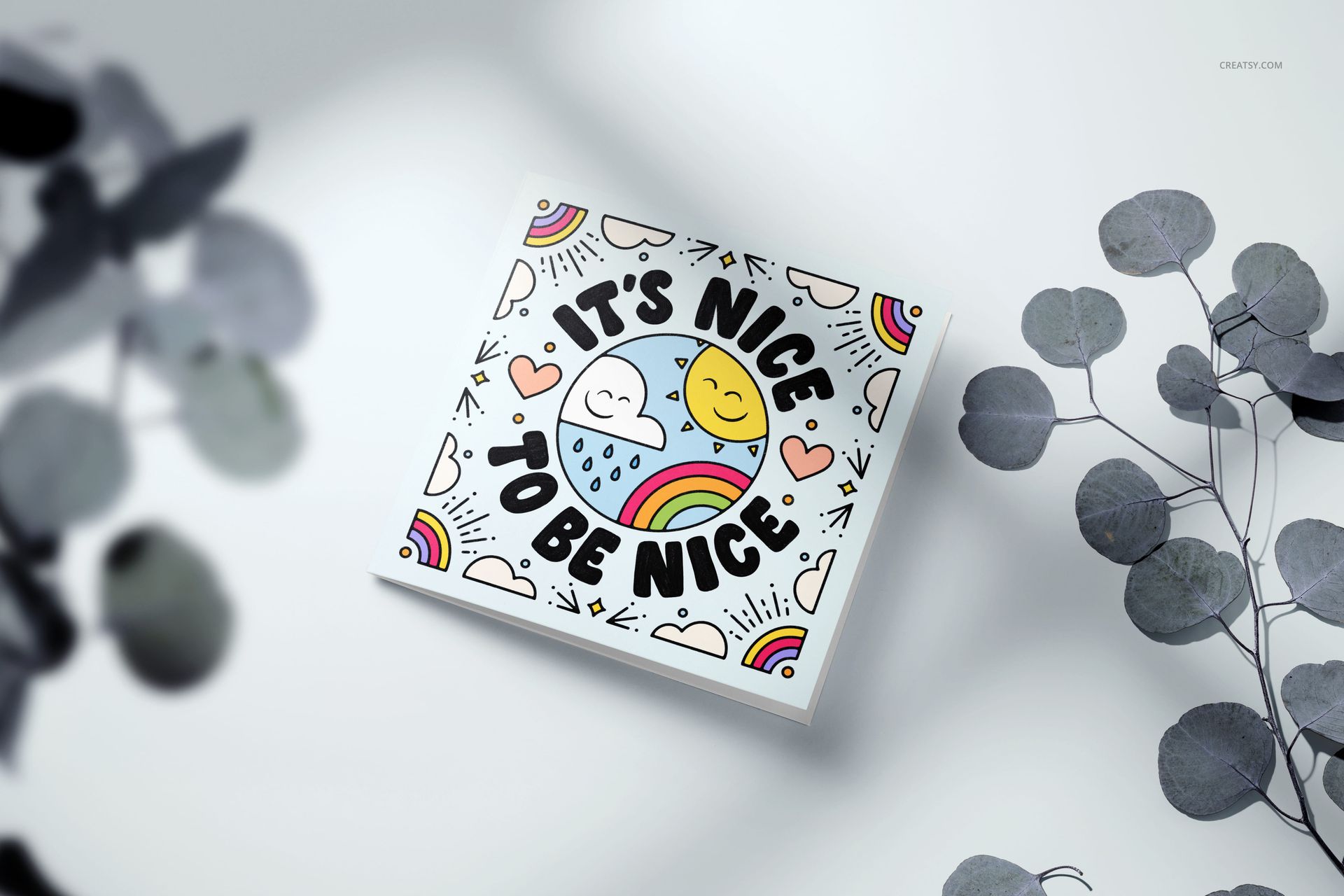 Square greeting card mockup featuring colorful artwork with rainbows, hearts, clouds, and a sun on a light surface.