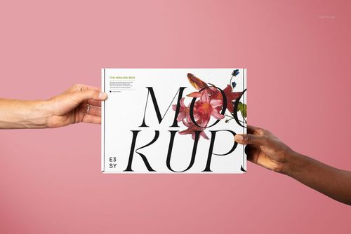 Mailing box mockup featuring large black letters and a flower graphic, held by two hands on a solid pink surface.