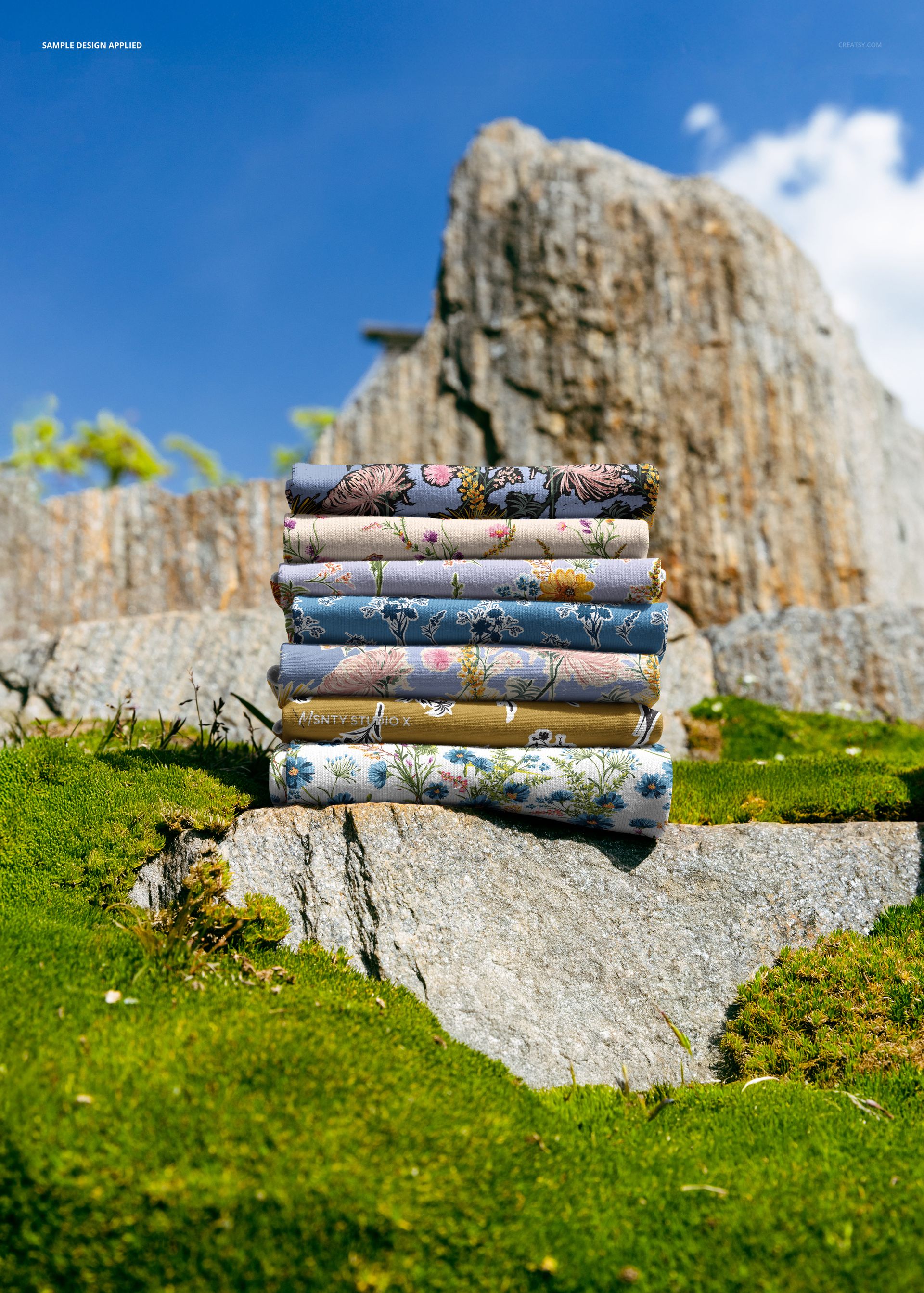 Stack of folded fabrics with floral patterns placed on a rocky outcrop, surrounded by green moss under a clear blue sky.