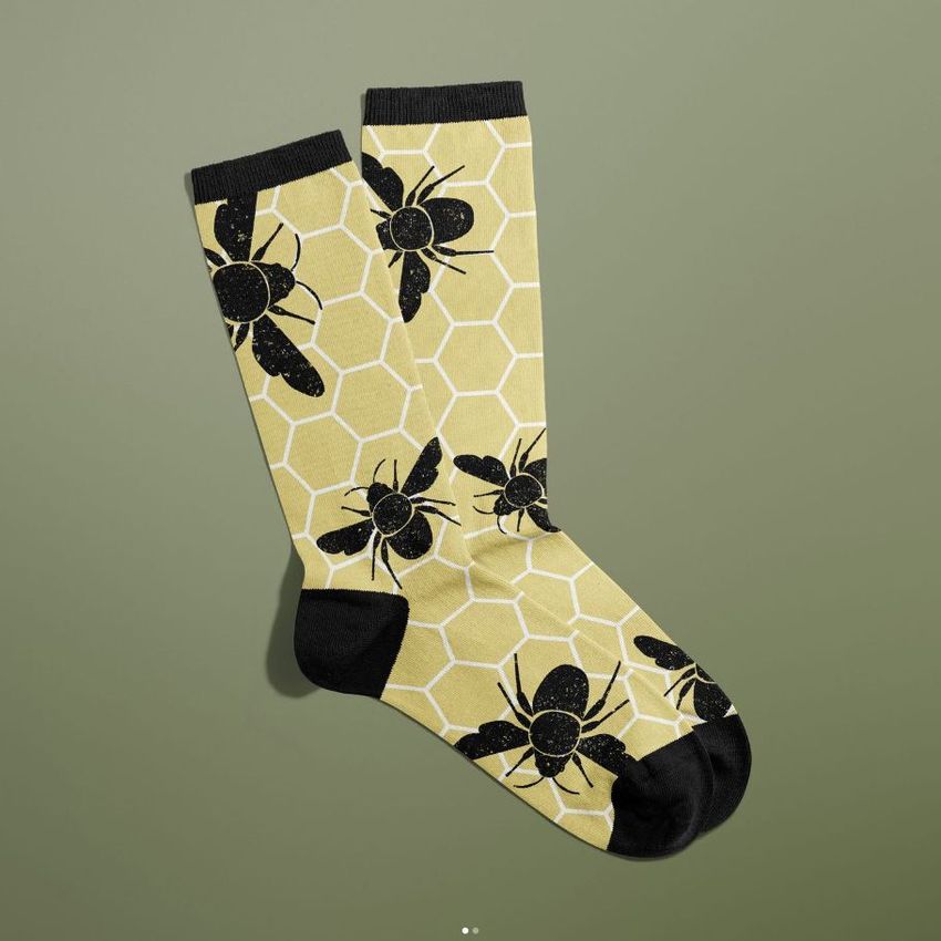 Socks mockup showing a pair of yellow socks with black bee illustrations and a honeycomb pattern, displayed on a green background.