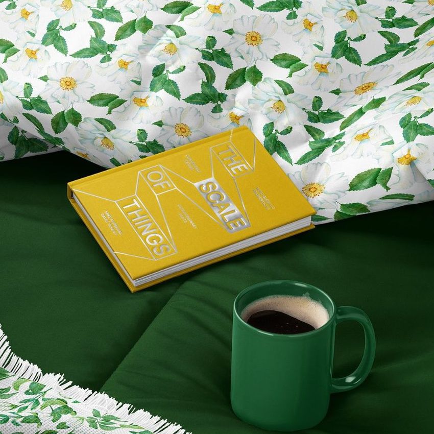 Microfiber comforter mockup featuring a floral pattern with white daisies and green leaves, a yellow book, and a green mug on a bed.