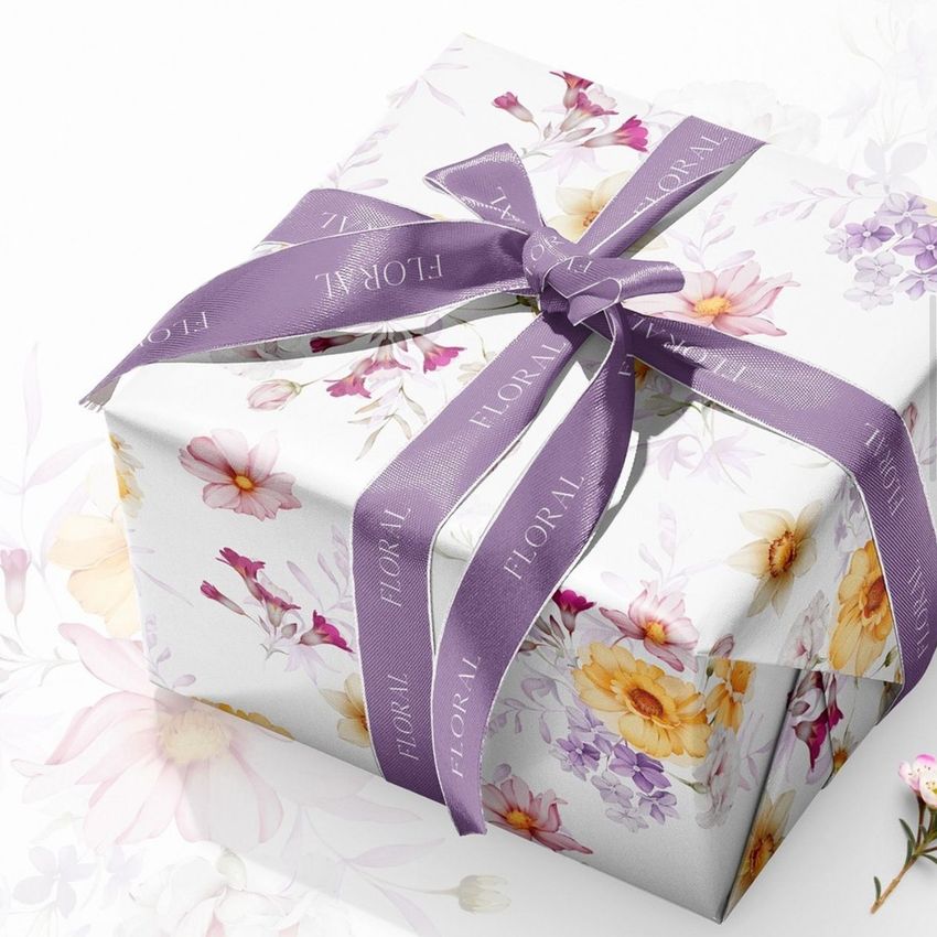 Gift box mockup wrapped in white paper with colorful floral patterns, tied with a purple ribbon featuring subtle text.