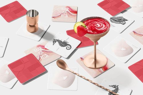 Square coasters in red and white displayed on a white surface with a cocktail glass, bar spoon, and copper cup.