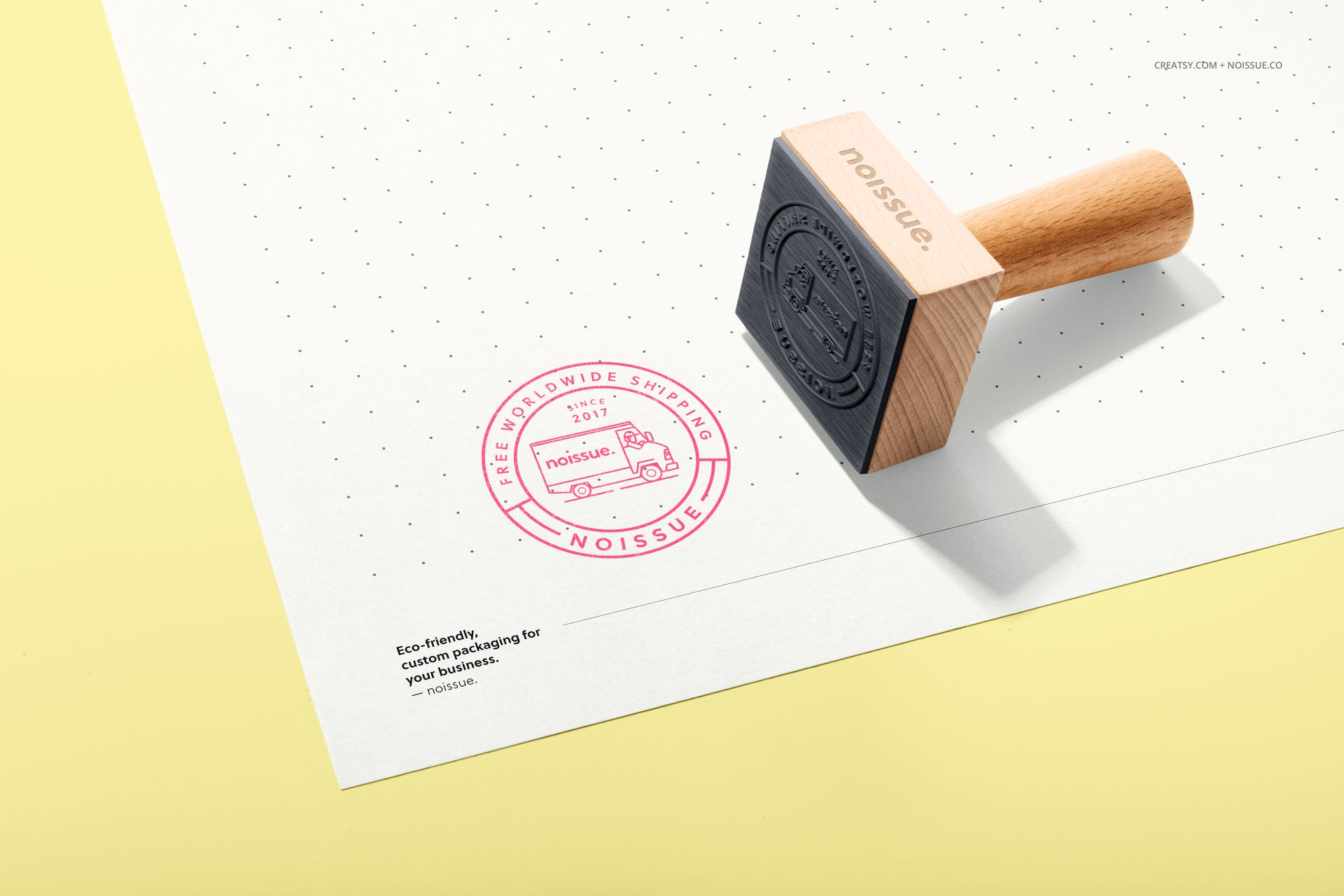 Noissue wooden stamp mockup displayed on white paper with dotted grid and pink ink stamp, angled top view.