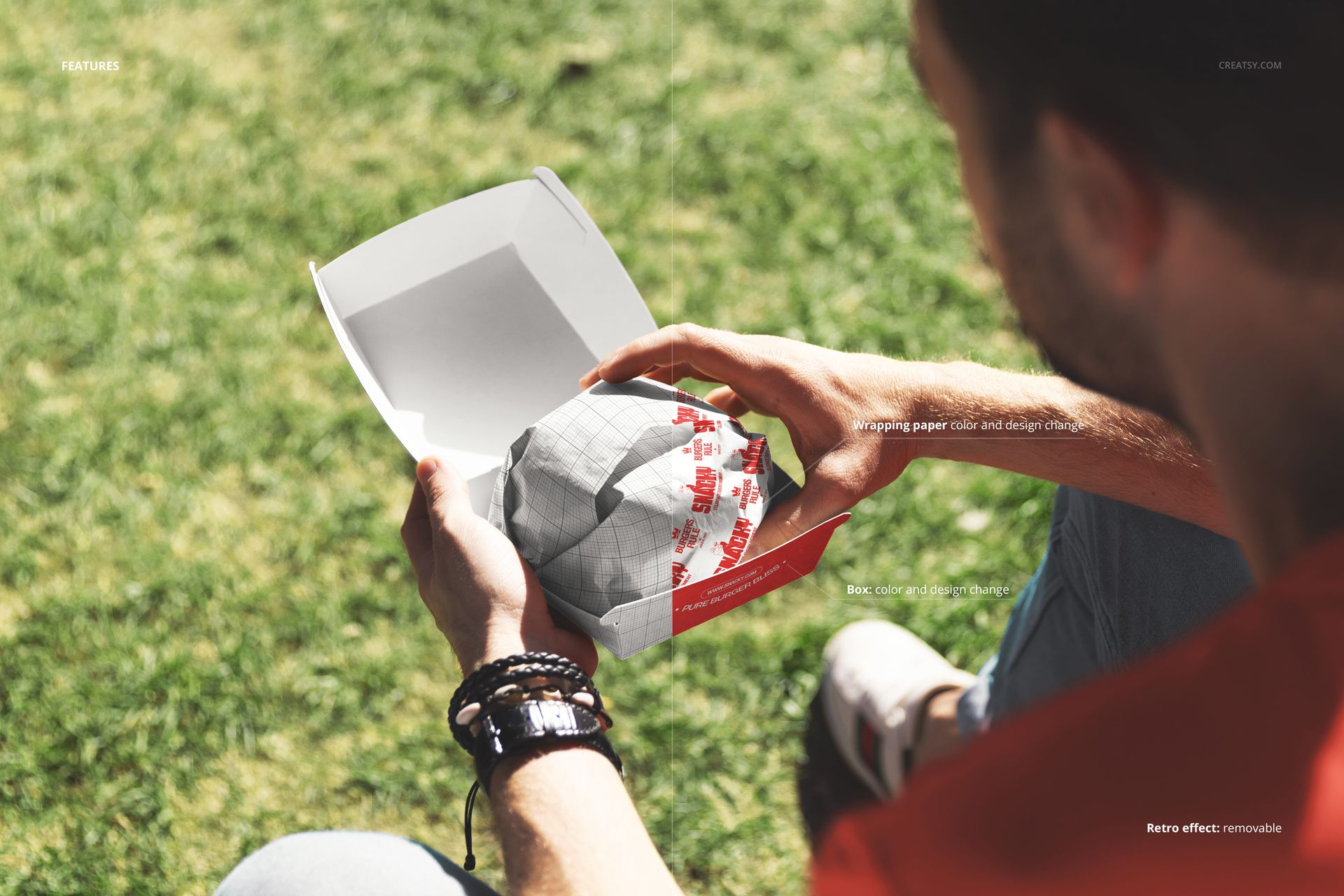 Person holding a wrapped burger in a white and red box outdoors on green grass, packaging mockup scene.