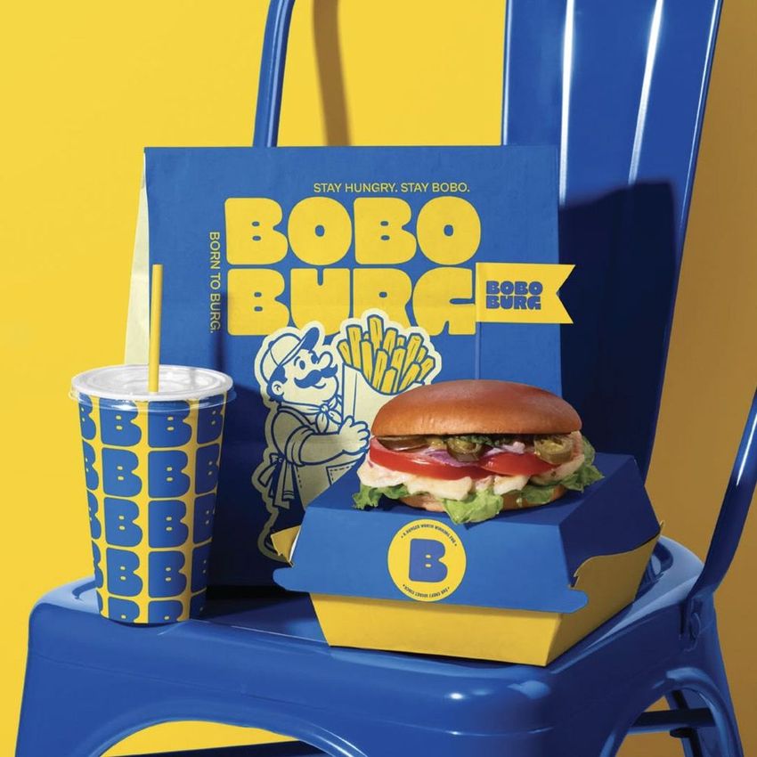 Restaurant branding mockup featuring a burger box, drink cup, and paper bag with bold blue and yellow graphics on a blue chair.