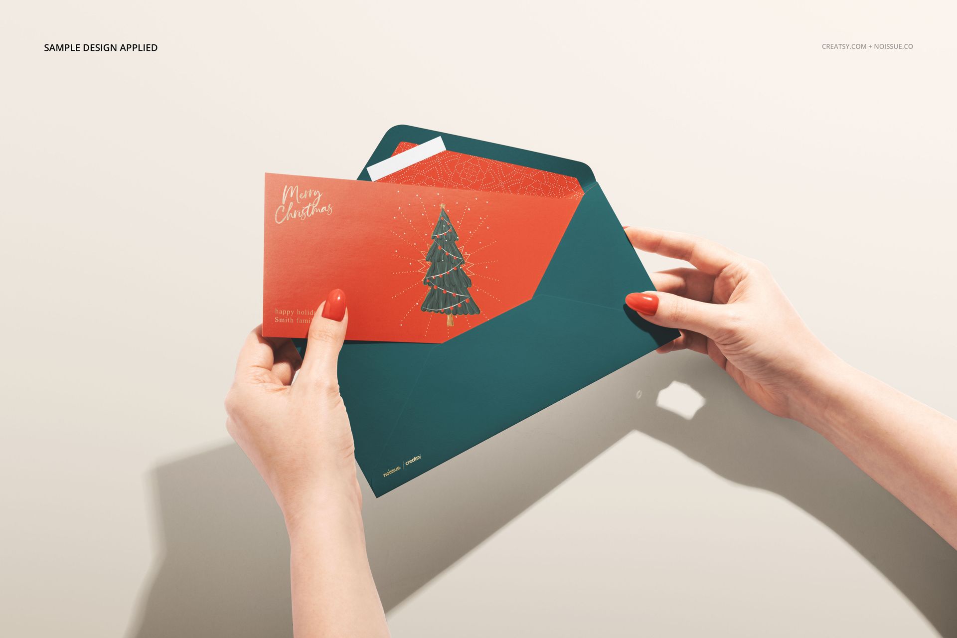 Person holding a stationery mockup set featuring a teal envelope and red greeting card with holiday design, top view.