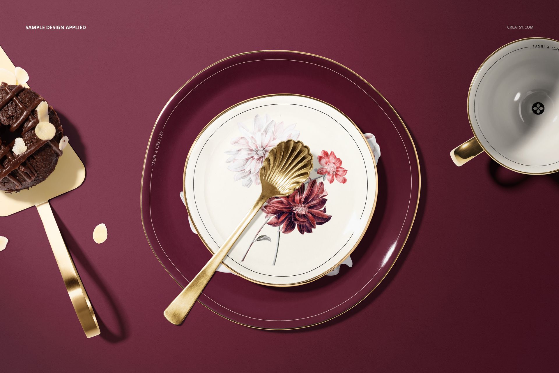 Top view of a plate mockup set with floral design, gold utensils, and a cup on a burgundy background.