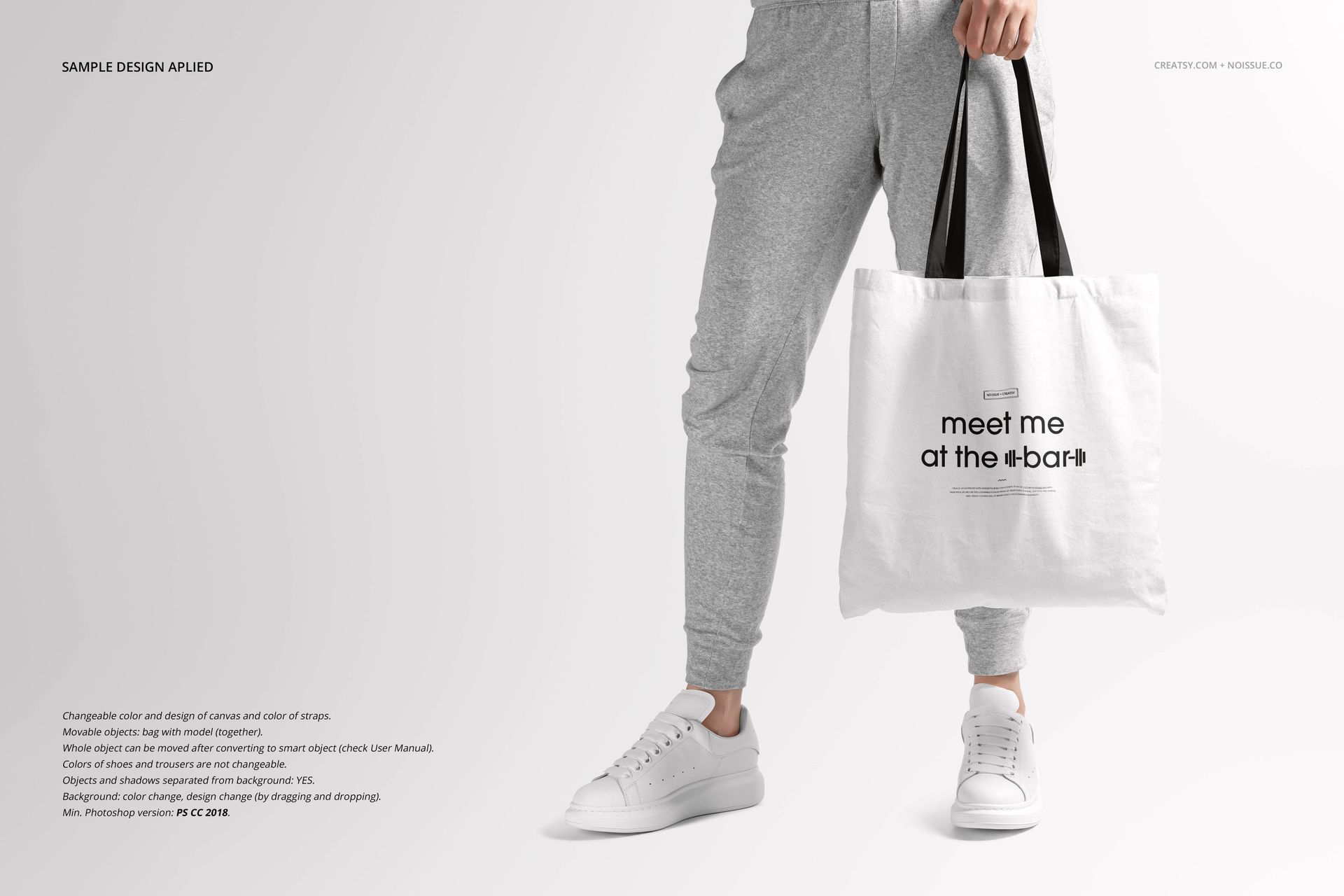 White tote bag mockup with black straps, displayed by a person wearing casual gray pants and white shoes.