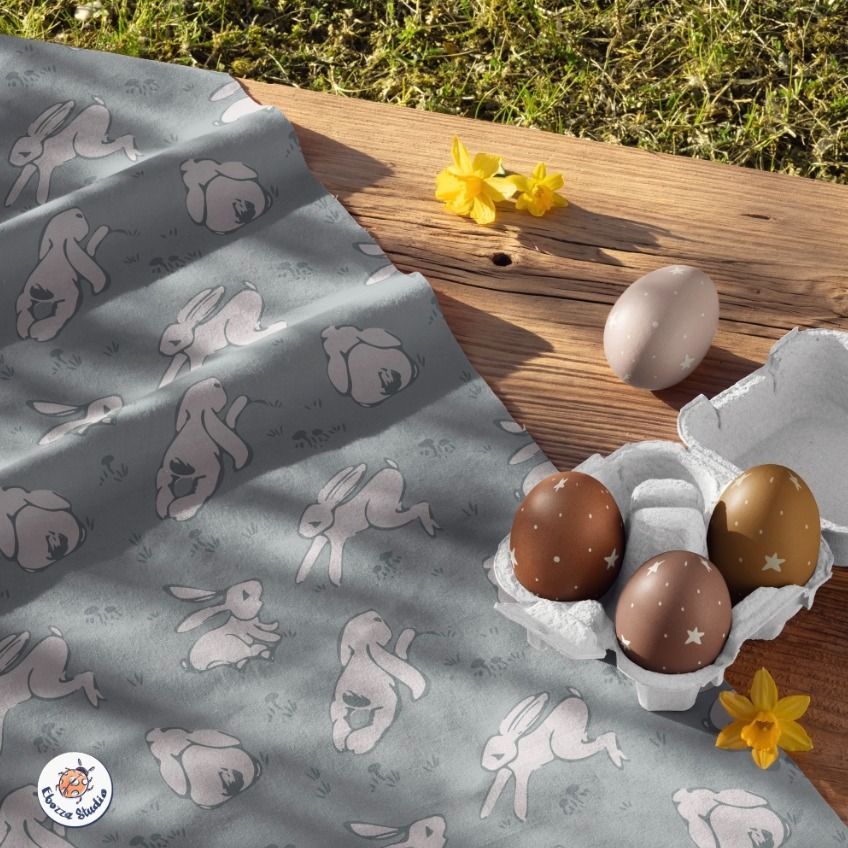 Cotton fabric mockup with whimsical bunny illustrations in soft pink on gray, displayed beside pastel Easter eggs on a wooden surface.