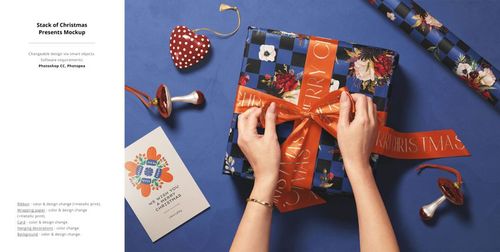 Hands tying an orange ribbon around a blue checkered gift box on a blue surface with Christmas wrapping props.