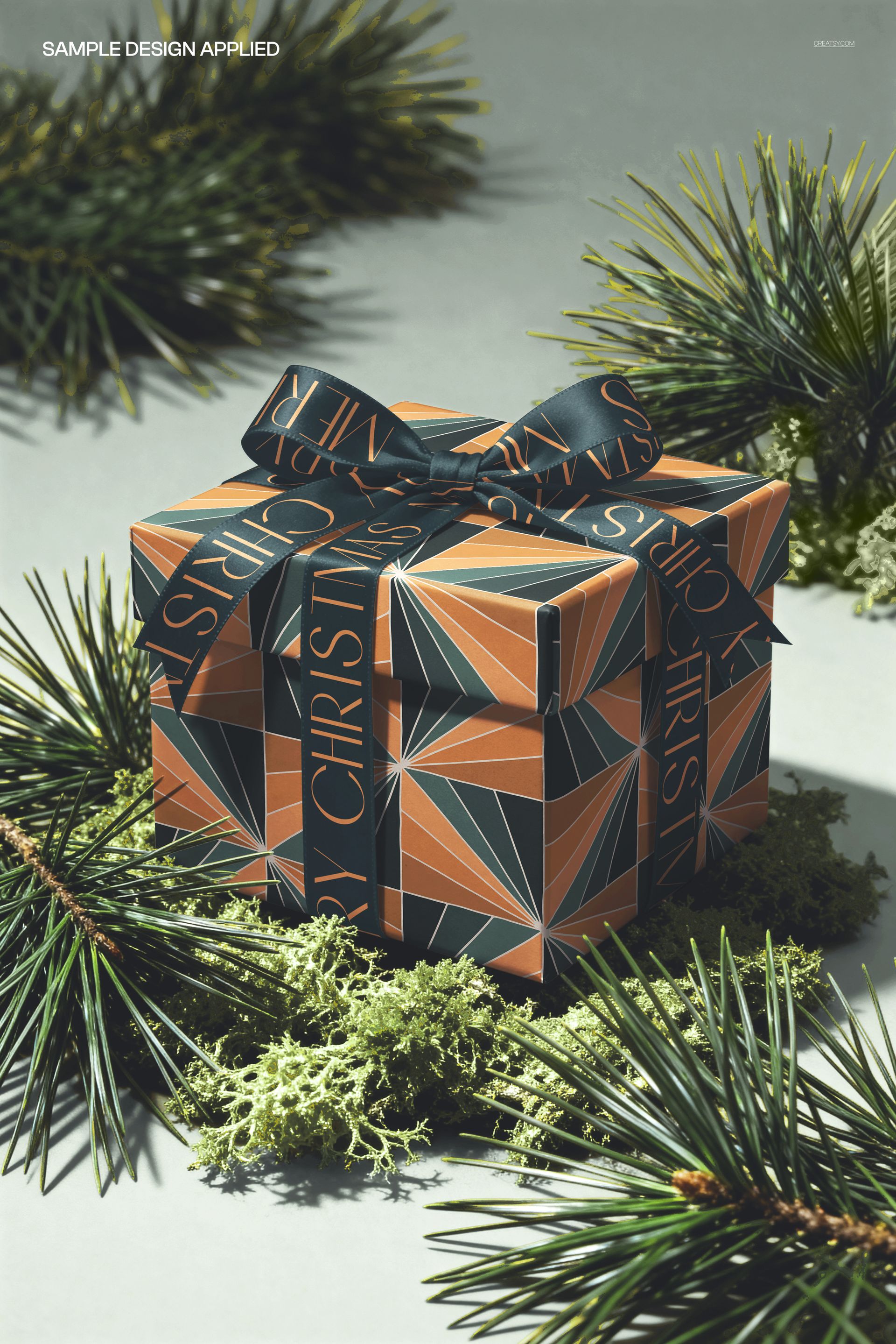 Front view of a square gift box mockup with a bow, featuring bold geometric design and conifers as props.