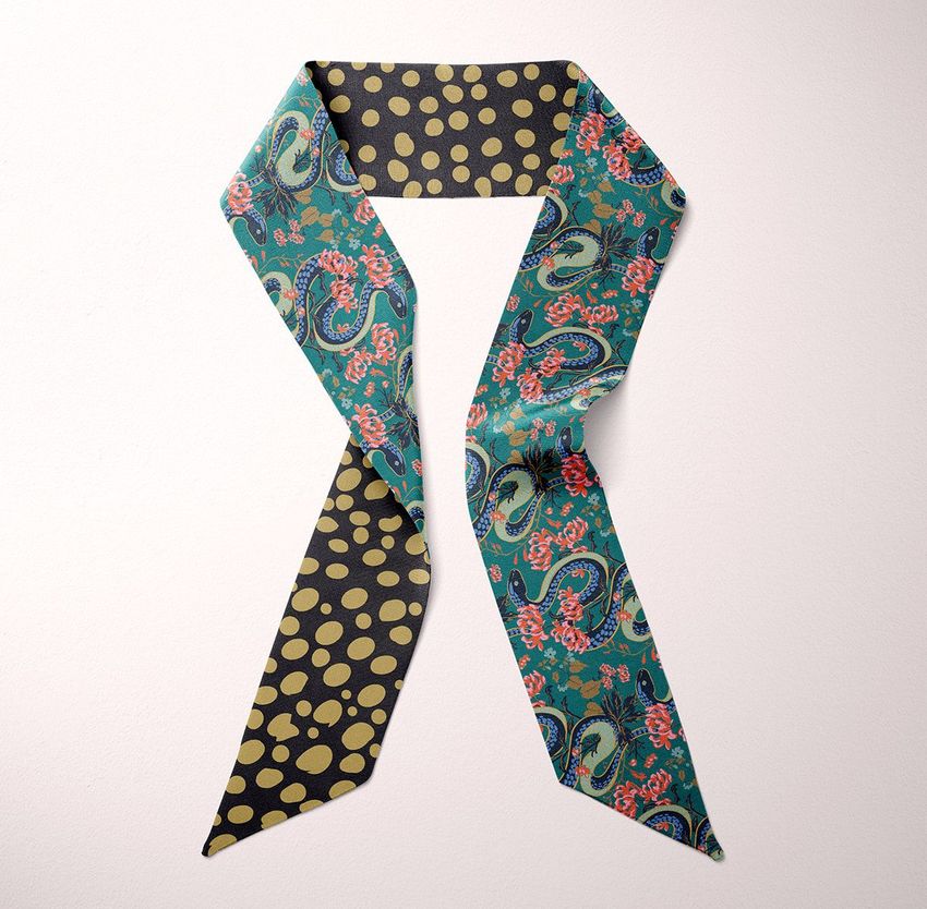 Top view of a twilly scarf mockup laid flat on a light surface, featuring a snake and floral pattern on one side and polka dots on the other.