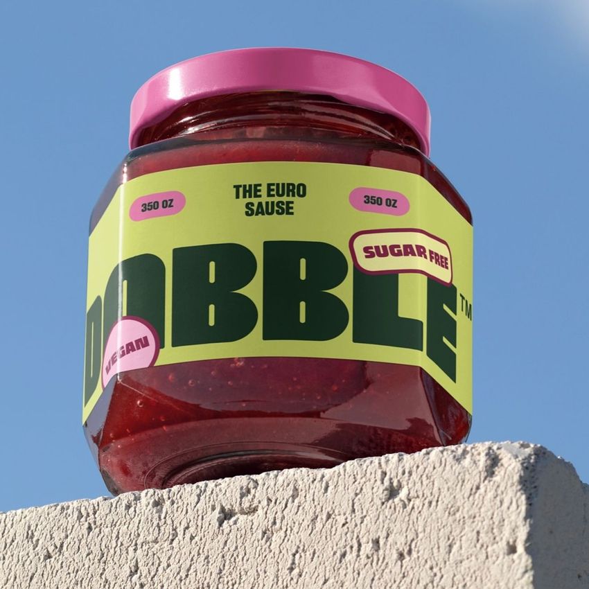 Raspberry jam jar mockup with a bold yellow and green label, pink lid, and visible text, placed on a textured concrete surface outdoors.