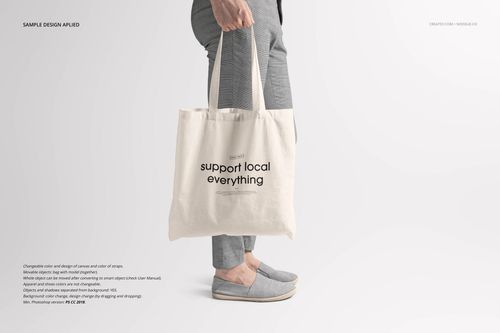 Front view of a canvas tote bag mockup held by a person wearing gray pants and slip-on shoes on a light background.