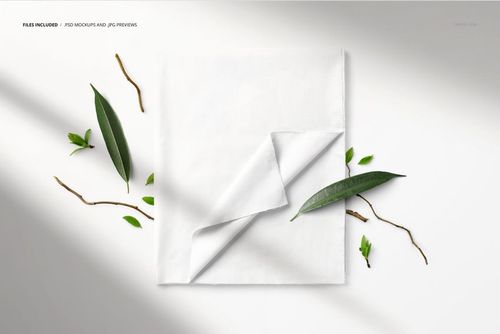 PSD mockup of a folded white textile surrounded by scattered green leaves and twigs, viewed from above.