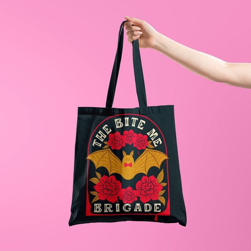 A black tote bag mockup held by a hand against a solid pink background, featuring a bat and red floral design with bold text.