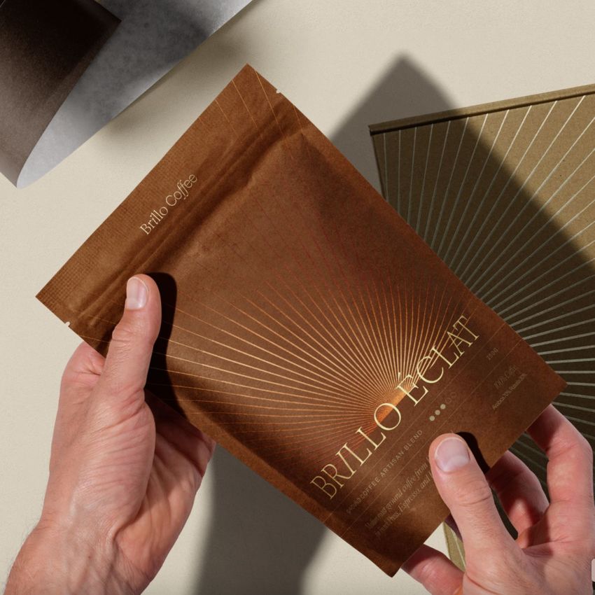 Hands holding a brown coffee packaging mockup with gold sunburst lines and metallic text on a light surface with other packaging nearby.