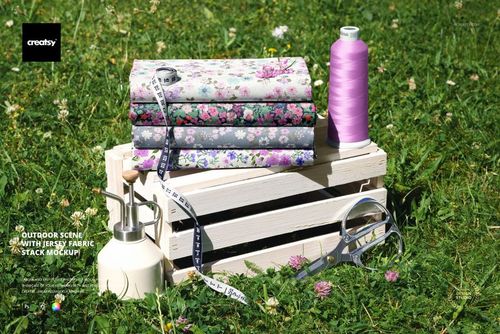 PSD mockup showing a pile of flower-print jersey fabrics on a white crate, with thread, tape measure, and scissors on grass.