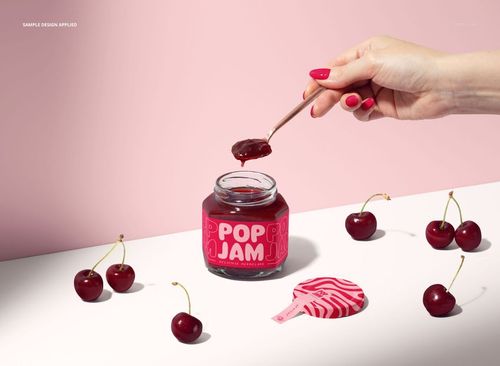 Hand holding a spoon above an open cherry jam jar mockup, with cherries and a pink lid on a pastel pink background.