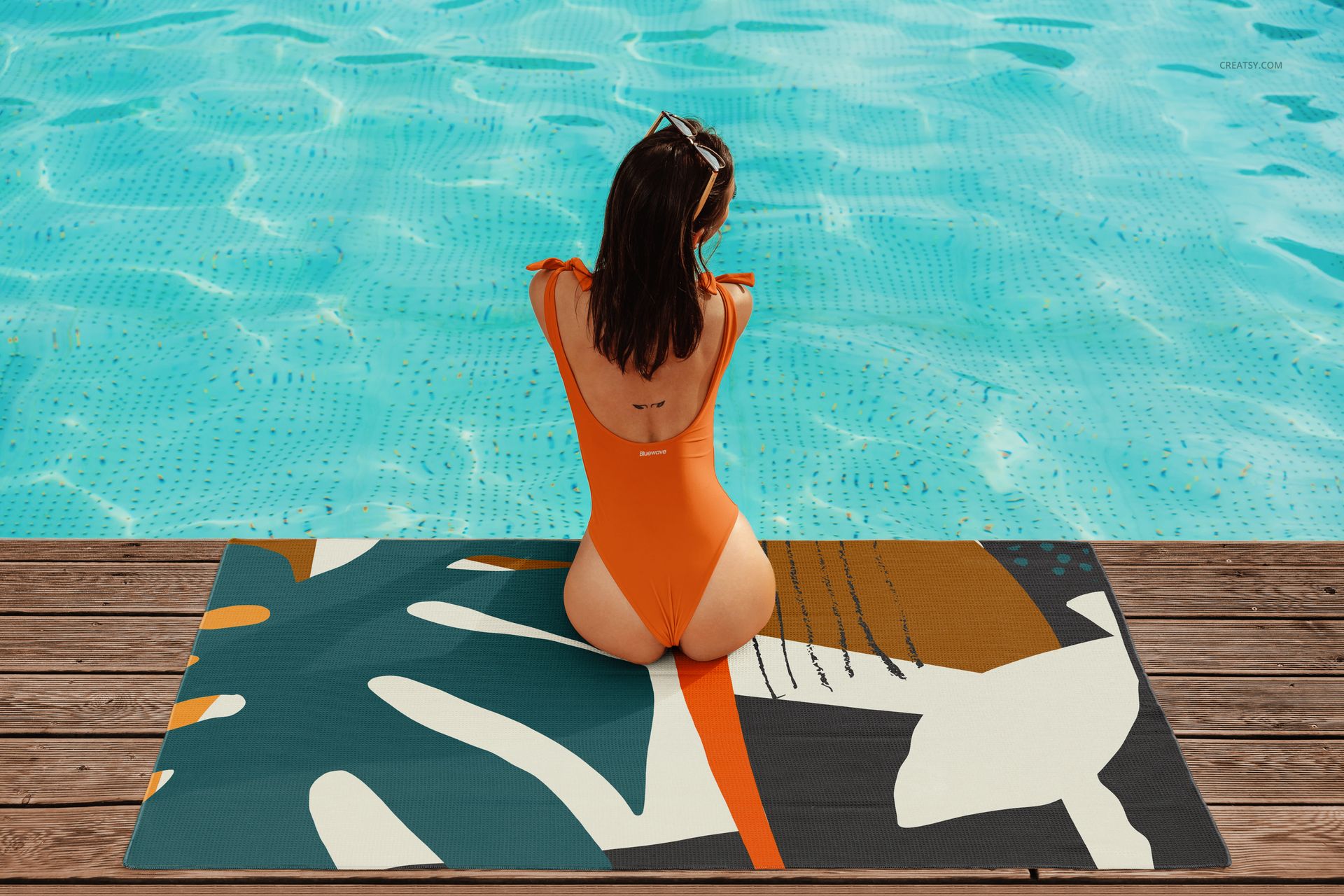 Colorful abstract beach waffle towel mockup with a woman in an orange swimsuit beside a clear blue swimming pool.