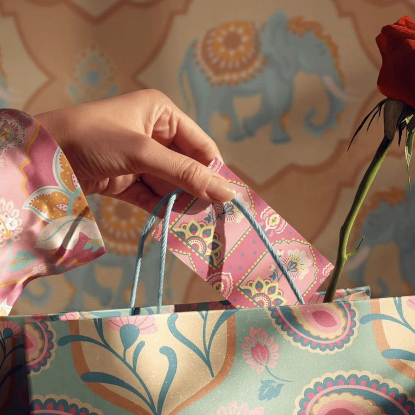 Hand placing a patterned card into a decorative gift bag with floral designs, next to a single red rose, with an elephant motif background.
