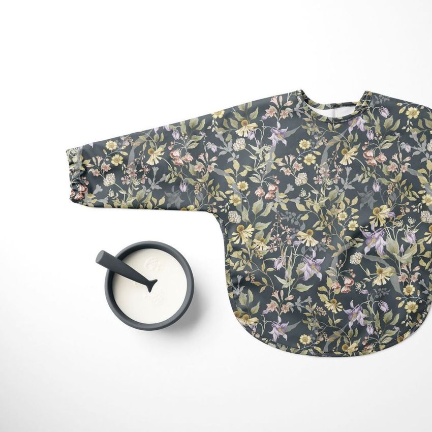 Baby sleeved bib mockup featuring a dark floral pattern, laid flat on a white surface next to a bowl of milk with a spoon.