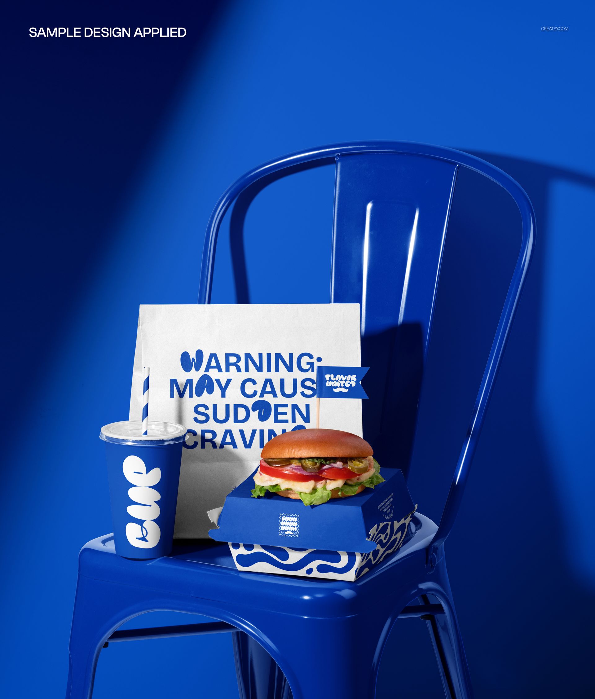Fast food packaging mockup featuring a paper bag, burger box, and drink cup on a blue metal chair in a studio setting.