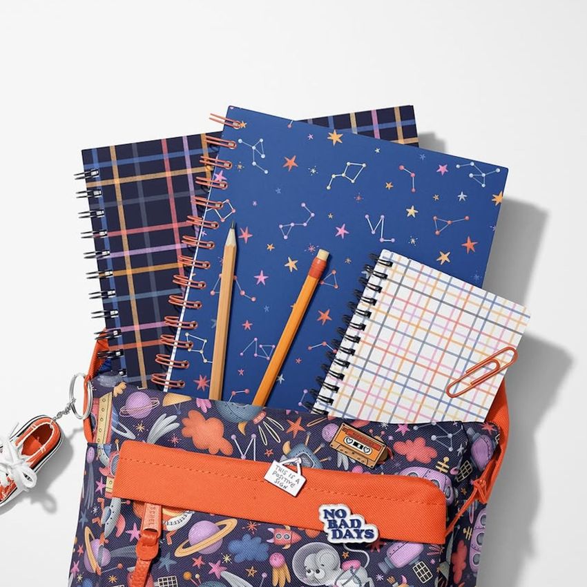 School backpack mockup with spiral notebooks in various patterns, two pencils, and a paperclip arranged on a white surface.