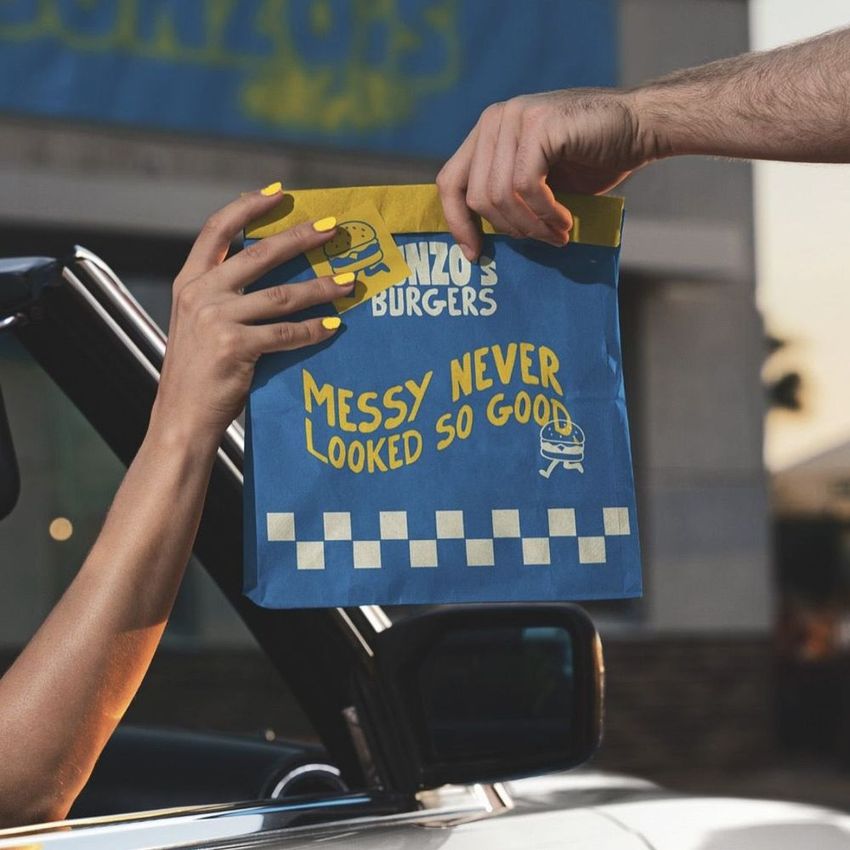 Fast food drive-thru bag mockup showing a blue and yellow paper bag being handed to a customer from a car window.