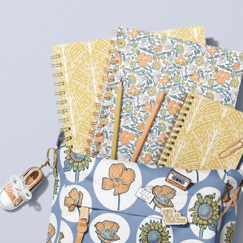 School backpack mockup with spiral notebooks in floral and leaf patterns, viewed from above on a light gray background.