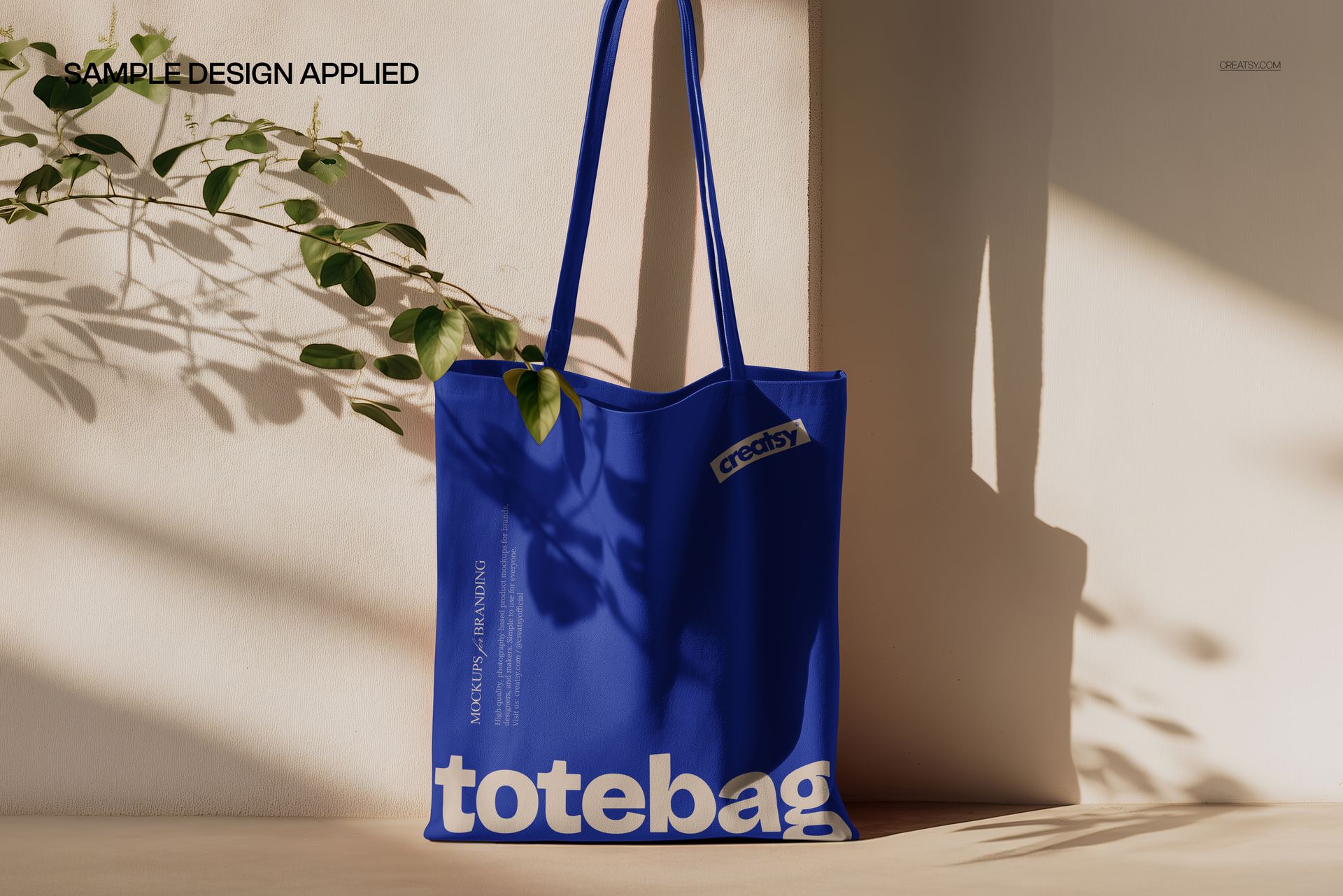 Blue canvas tote bag mockup hanging against a light wall, with plant shadows cast across the surface.