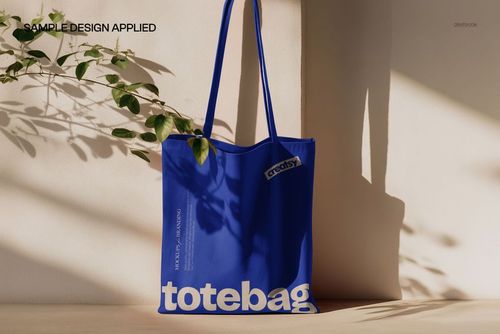 Blue canvas tote bag mockup hanging against a light wall, with plant shadows cast across the surface.
