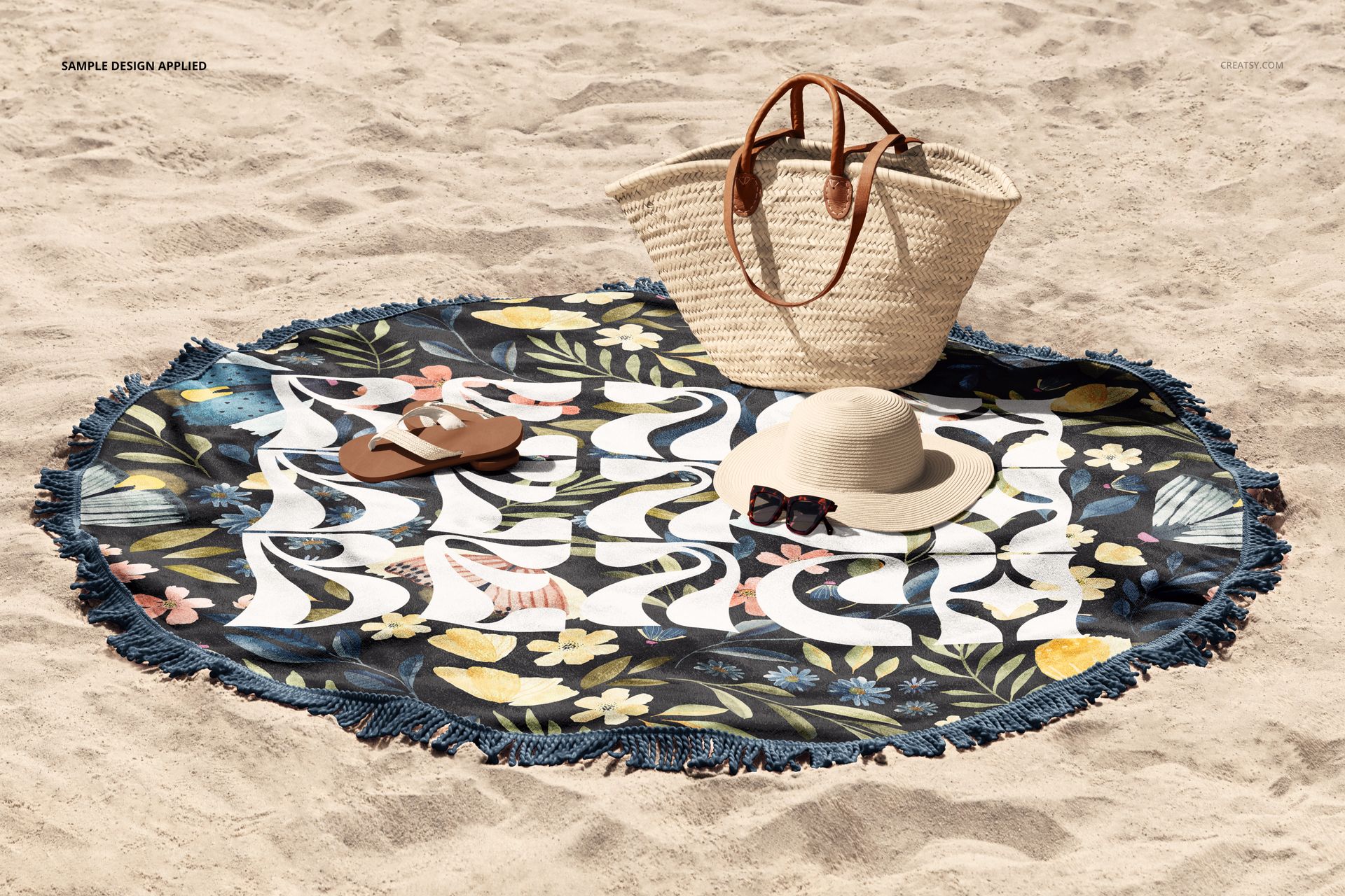 Top view of a round beach towel mockup featuring blue fringe, floral design, and props like hat and bag on sand.
