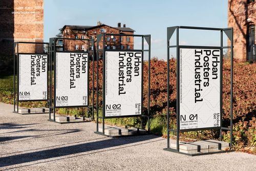 Urban posters mockup set shown in metal stands, positioned in front of industrial buildings and landscaped bushes.